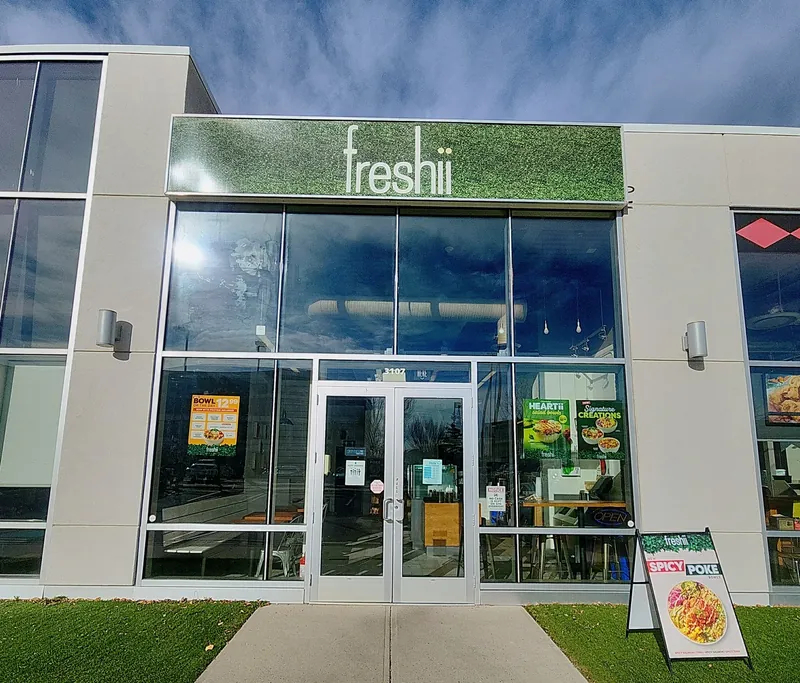 Front entrance of Freshii Sundance in Sunpark Plaza, SE Calgary, with the Spicy Poke Bowls sidewalk sign and unit number 3107 above the doors.